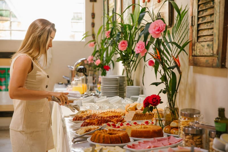 Hóspede se servindo no buffet de café da manhã com bolos caseiros, pães e flores